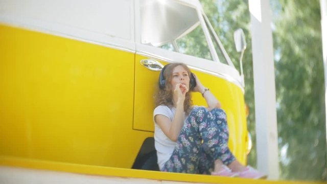young woman outdoors by yellow vagon car listening music in headphones using smartphone - relaxing, enjoying, concept of technology and travel