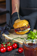 Tasty beef burger hold in the hands grilled with lettuce and mayonnaise served with potato fries and fresh vegetables on rustic wooden table on dark background