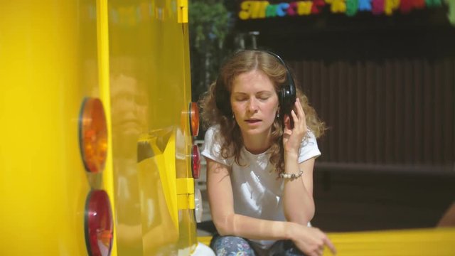 young woman outdoors by yellow vagon car listening music in headphones using smartphone - relaxing, enjoying, concept of technology and travel