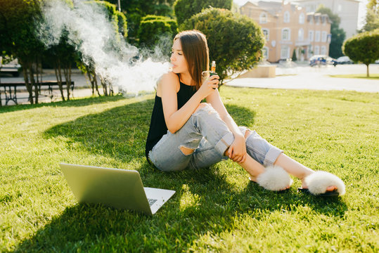 Outdoors photo of trendy attractive girl smoking vape, spending leisure time while sitting on grass in the park, with laptop.