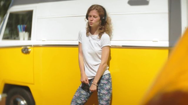 young woman outdoors by yellow vagon car listening music in headphones using smartphone - relaxing, enjoying, concept of technology and travel