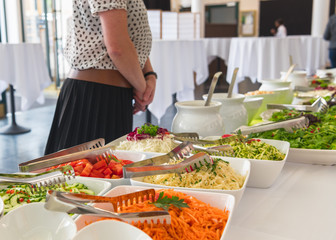 Frau steht vor einem Catering Buffet mit frischen vegetarischen Salaten in Schalen zum selberbedienen mit Tellern und Soßen