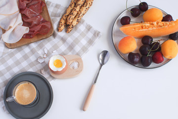 Perfectly served breakfast boiled egg in wooden egg cup with breadsticks ham melon cherry apricots coffee salt spoon and checkered napkin White background Top view Copy space