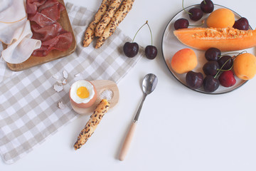 Perfectly served breakfast boiled egg in wooden egg cup with breadsticks ham melon cherry apricots salt spoon and checkered napkin White background Top view Copy space