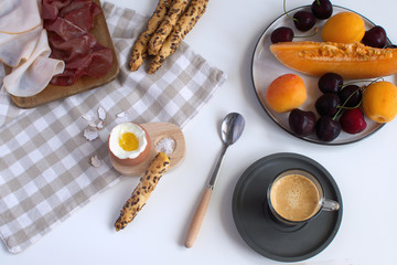Perfectly served breakfast boiled egg in wooden egg cup with breadsticks ham melon cherry apricots coffee salt spoon and checkered napkin White background Top view Copy space