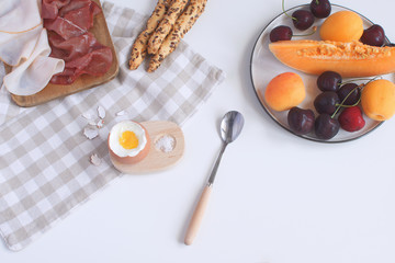 Perfectly served breakfast boiled egg in wooden egg cup with breadsticks ham melon cherry apricots salt spoon and checkered napkin White background Top view Copy space