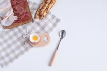 Perfectly served breakfast boiled egg in wooden egg cup with breadsticks ham melon cherry apricots salt spoon and checkered napkin White background Top view Copy space