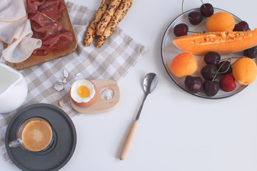 Perfectly served breakfast boiled egg in wooden egg cup with breadsticks ham melon cherry apricots coffee salt spoon and checkered napkin White background Top view Copy space