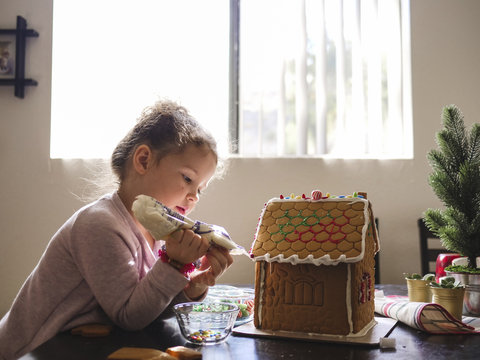 Side view of girl icing on gingerbread house while sitting at home&dagger;