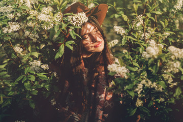 beautiful hipster girl in hat  posing in green leaves with flowers in evening sunlight among shadows. stylish woman traveler calm portrait in spring garden. space for text. atmospheric moment