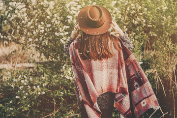 stylish hipster girl in hat and poncho with beautiful hair with flowers walking in park in evening...