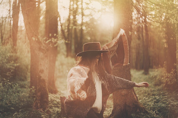Fototapeta na wymiar stylish hipster traveler girl in hat exploring in woods in amazing evening sunshine light. woman having fun and jumping in sunlight. space for text. atmospheric moment. wanderlust