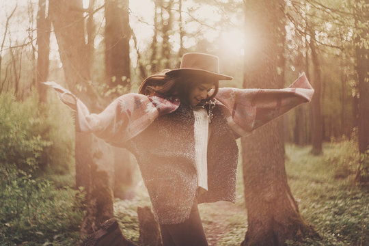 Stylish Hipster Traveler Girl In Hat Exploring In Woods In Amazing Evening Sunshine Light. Woman Having Fun And Jumping In Sunlight. Space For Text. Atmospheric Moment. Wanderlust