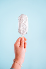 Woman's hand holding ice-cream, popsicle wrapped in silver foil. Pastel blue background. Minimalism food photography. Copyspace