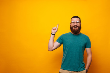 Happy smiling young bearded man wearing glasses and pointing up over yellow background