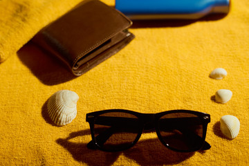Wallet, sunglasses, sun cream and sea shells on yellow towel in sunlight. Copy space for text. 