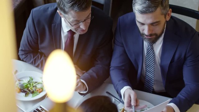 High angle view of two businessmen in suits and neckties sitting at table at lunch and negotiating contract with unrecognizable colleague