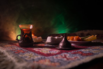 Arabian tea in glass with eastern snacks on a carpet on dark background with lights and smoke. Eastern tea concept. Empty space.