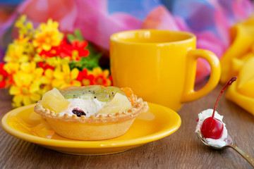 Tea and dessert in yellow colors. A yellow cup of tea and a bright cake with a cherry on a background of colorful fabric and flowers. Tea and dessert in the process of eating.