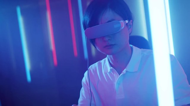 East Asian Pro Gamer Puts On Virtual Reality Headset Plays Online Video Game with Joysticks / Controllers. Cool Retro Neon Colors in the Room.
