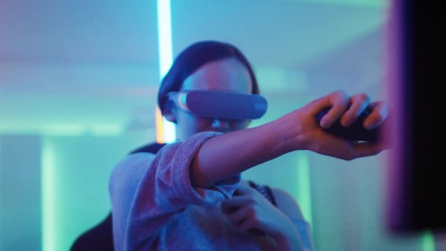 Pro Gamer Girl Wearing Virtual Reality Headset Plays Sword Fighting Online Video Game with Joysticks / Controllers. Cool Retro Neon Colors in the Room.