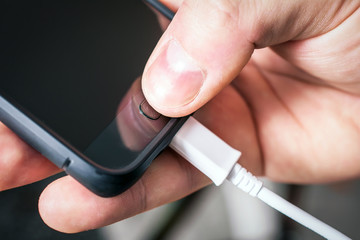 Using A Black Mobile Phone, Connected To A White USB Cable, Holded By A Male Hand