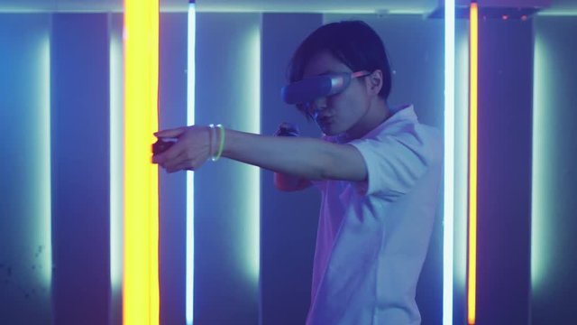 East Asian Pro Gamer Wearing Virtual Reality Headset Plays Online Video Game Shooter using Joysticks / Controllers as Shotgun / Gun. Cool Retro Neon Colors in the Room.
