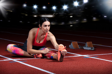 young athletic woman doing exercises on flexibility. stretching legs sitting track stadium, night with reflectors - obrazy, fototapety, plakaty
