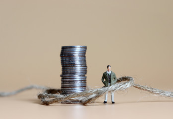 A miniature man connected with a pile of coins and rope.