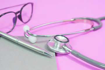 Stethoscope with glasses on blank notebook and pen on pink background.