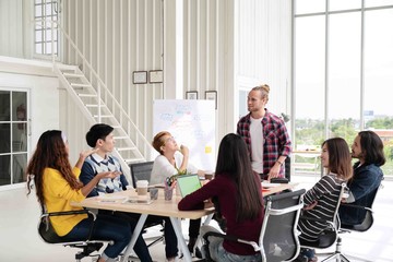 Obraz premium Young caucasian creative man standing and making presentation at modern office happy talking and brainstorming with team. Casual multiethnic diverse people business meeting concept with rear view.
