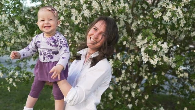 Happy caucasian 20s mum play with pretty adorable small infant outside. Mommy hold, looking, play, hugging near with flowers tree at background at spring time in windy day close up. Two beauty laught