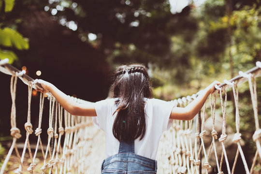 Cute Asian Child Girl Walking On The Hanging Rope Bridge Across The River In The Forest With Fun