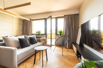 Elegant gray sofa and a big flat screen TV in a modern living room interior with hardwood herringbone floor and open balcony glass door