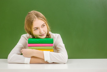 Teen girl hugs a stack of books on the background of a school board and looking away. Space for text