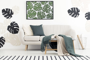 Green blanket on stool in front of couch in living room interior with poster of leaves. Real photo