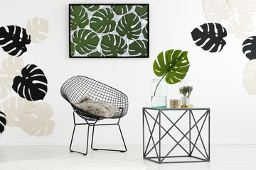 Table next to black armchair in white living room interior with poster of monstera leaves. Real photo