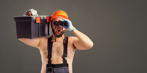 Funny fat bearded builder with binoculars on a gray background.