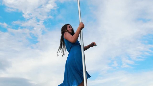 Attractive woman in blue dress performing pole-dance against the sky