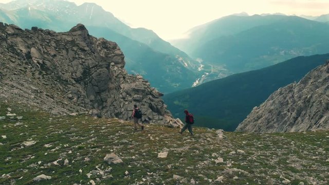 Aerial: couple backpackers hiking to the mountain top, rocky mountain peak in backlight, scenic landscape. Summer adventures on the Alps. Slow motion. Conquering success leader concept