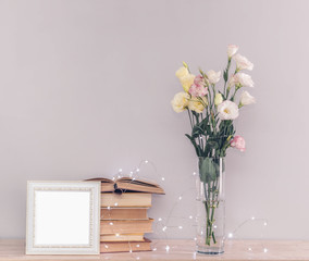 Eustoma flowers bouquet in a vase, stack of old vintage books, white photo frame and garland lights on a grey background. Reading and relax concept.
