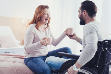 Obraz premium Funny joke. Positive optimistic disabled man drinking tea and looking at his emotional girlfriend laughing at his funny joke