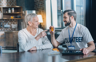 Healthcare. Good-looking exuberant bearded man smiling and holding some pills while talking with an old man