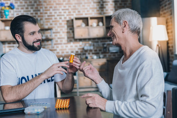 Amazing pills. Handsome joyful dark-eyed man smiling and holding some pills while talking with an old man