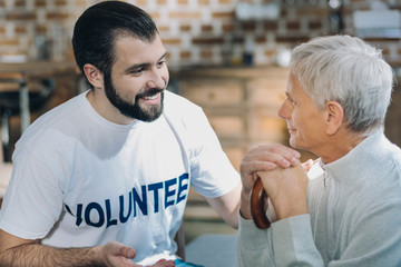 I trust you. Attractive cheerful bearded volunteer smiling and talking with an old man while sitting in the kitchen