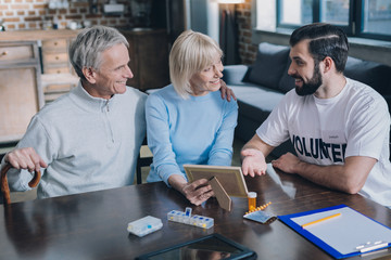 Cheerful aged spouses smiling and showing photos to a young man