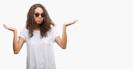 Young hispanic woman wearing sunglasses clueless and confused expression with arms and hands...