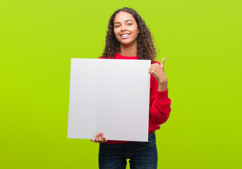 Young hispanic woman holding blank banner happy with big smile doing ok sign, thumb up with fingers, excellent sign