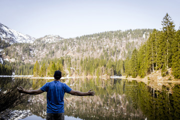 Fototapeta na wymiar young man with cap and blue shirt is standing in front of mountain lake with raising arms