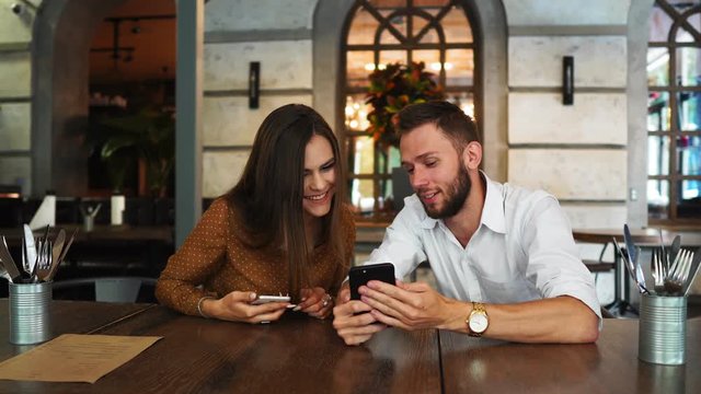Best female and male friends watch funny video on cell phone, rest together at outdoor cafe, drink cocktail, being in high spitrit. Cheerful tourists view photos on cellular, connected to internet
